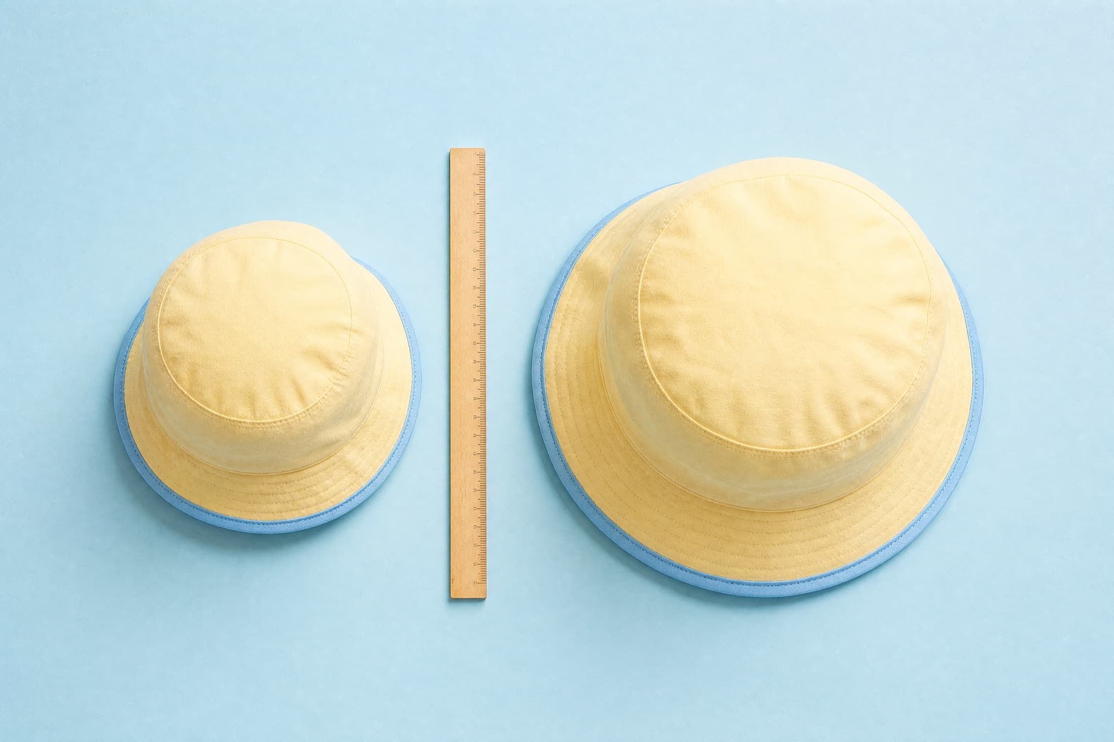 A child-size and an adult-size butter-yellow bucket hat laid on a sky-blue surface with a wooden ruler standing between them